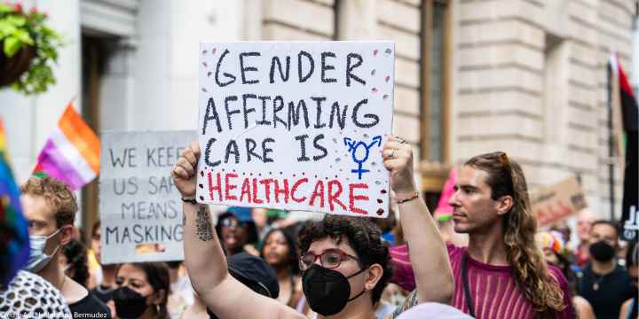 A shirtless demonstrator (wearing glasses and a black surgical mask) holds up a sign that reads" GENDER AFFIRMING CARE IS HEALTHCARE".